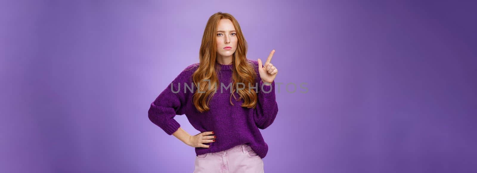 I am watching you. Portrait of bossy and suspicious cute redhead sister in purple sweater squinting doubtful and serious as making assumptions or hesitating pointing at upper left corner.