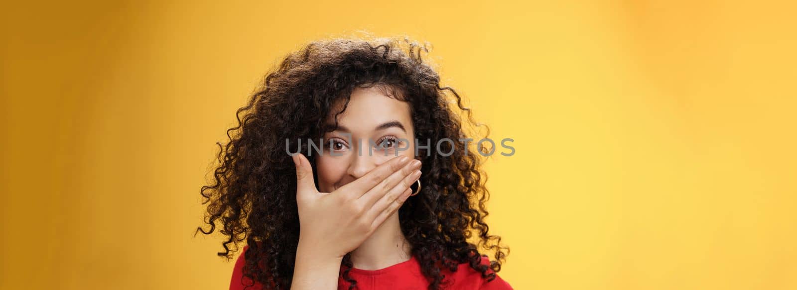 Close-up shot of excited and happy attractive female with curly hair giggling, chuckling and covering mouth as smiling standing amused and joyful as mocking friend over yellow background by Benzoix