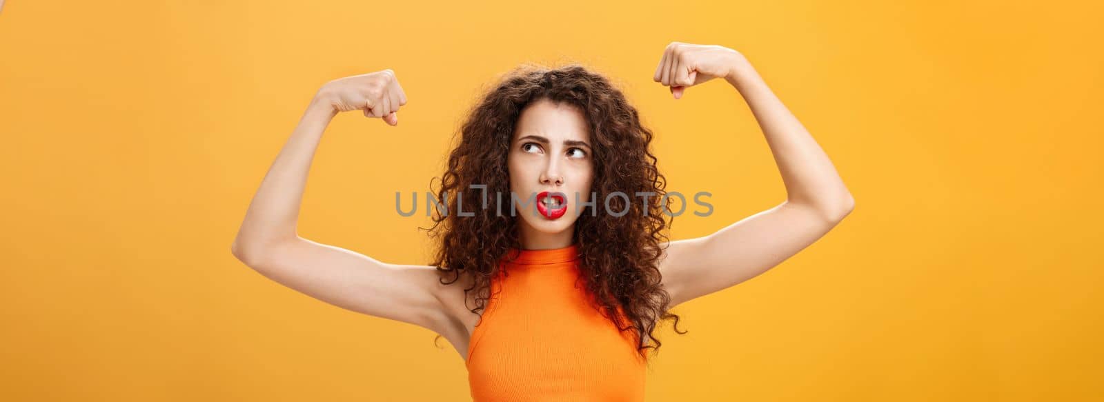 Woman feeling powerful and strong raising hands. with clenched fists making intense face being working out in gym showing muscles and biceps looking at upper right corner posing over orange background.