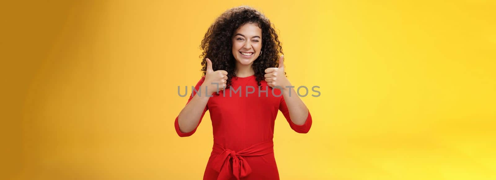 Creative and charismatic happy upbeat woman 25s with curly hair in red dress winking in approval and showing thumbs up with broad smile, satisfied giving positive reply over yellow wall by Benzoix