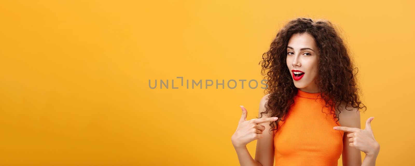 Proud and satisfied cool urban female with red lipstick. and curly hairstyle pointing at herself with self-assured expression winking bragging about skills and achievements over orange background.