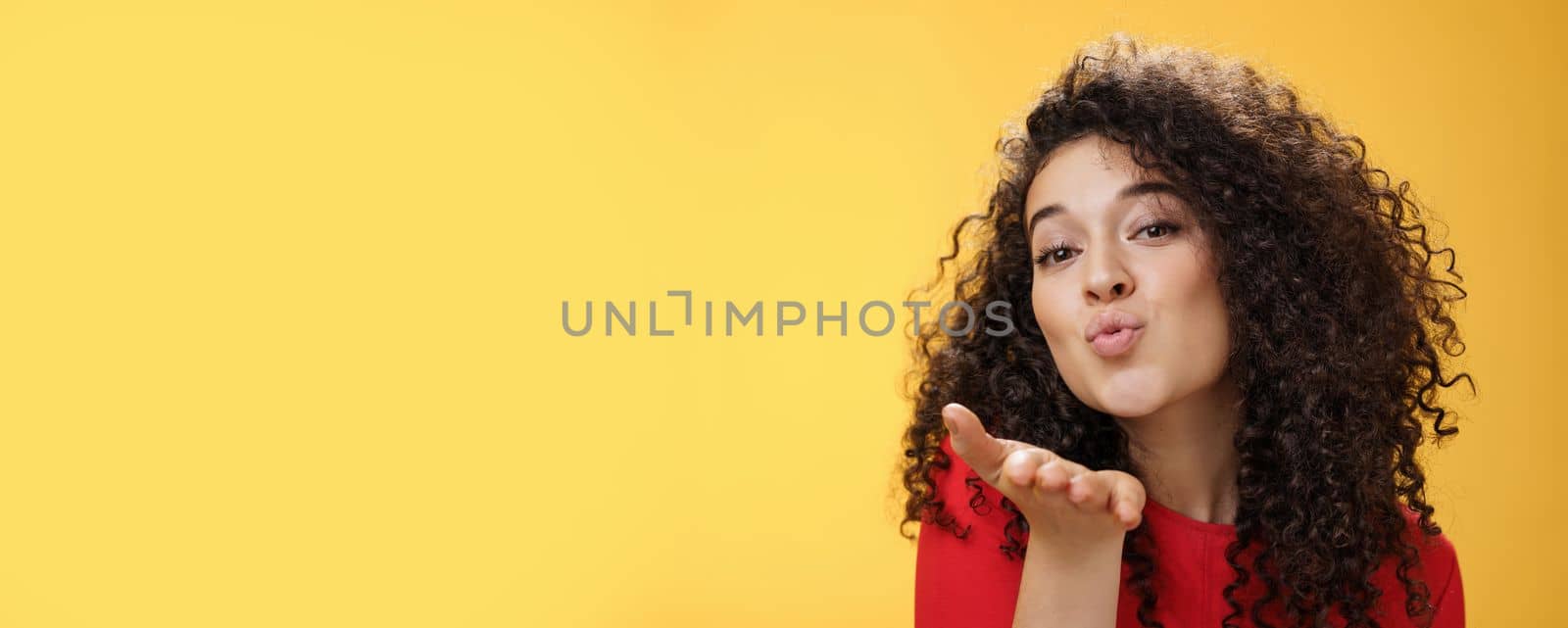 Sensual flirty and tender attractive girlfriend with curly hairstyle folding lips extending hand to send air kiss at camera, smiling flirting and being coquettish over yellow background by Benzoix