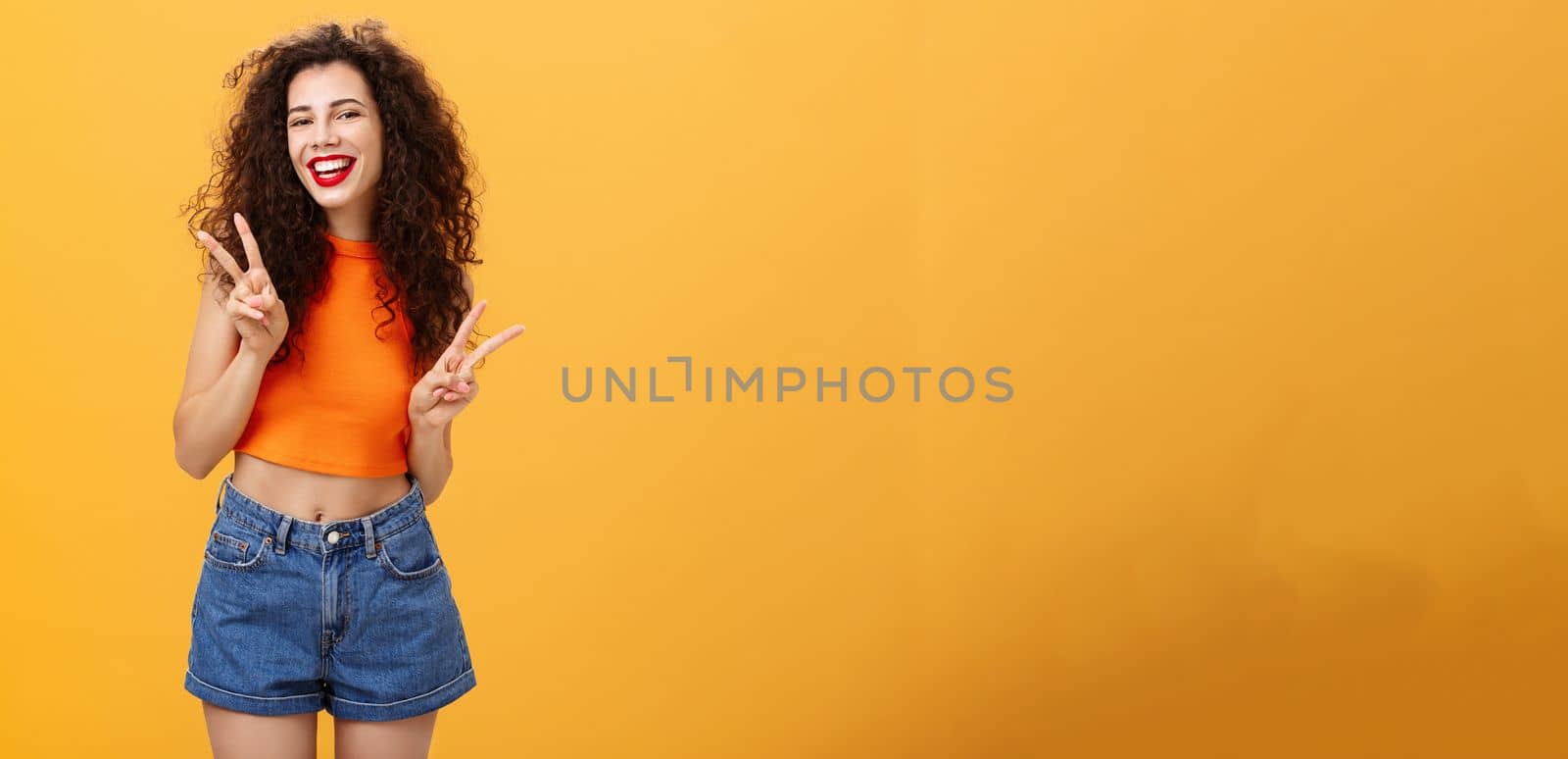 Outgoing friendly-looking attractive young female. with curly haircut in cropped top and shorts showing peace or victory signs with both hands and smiling cheerfully having fun over orange background.