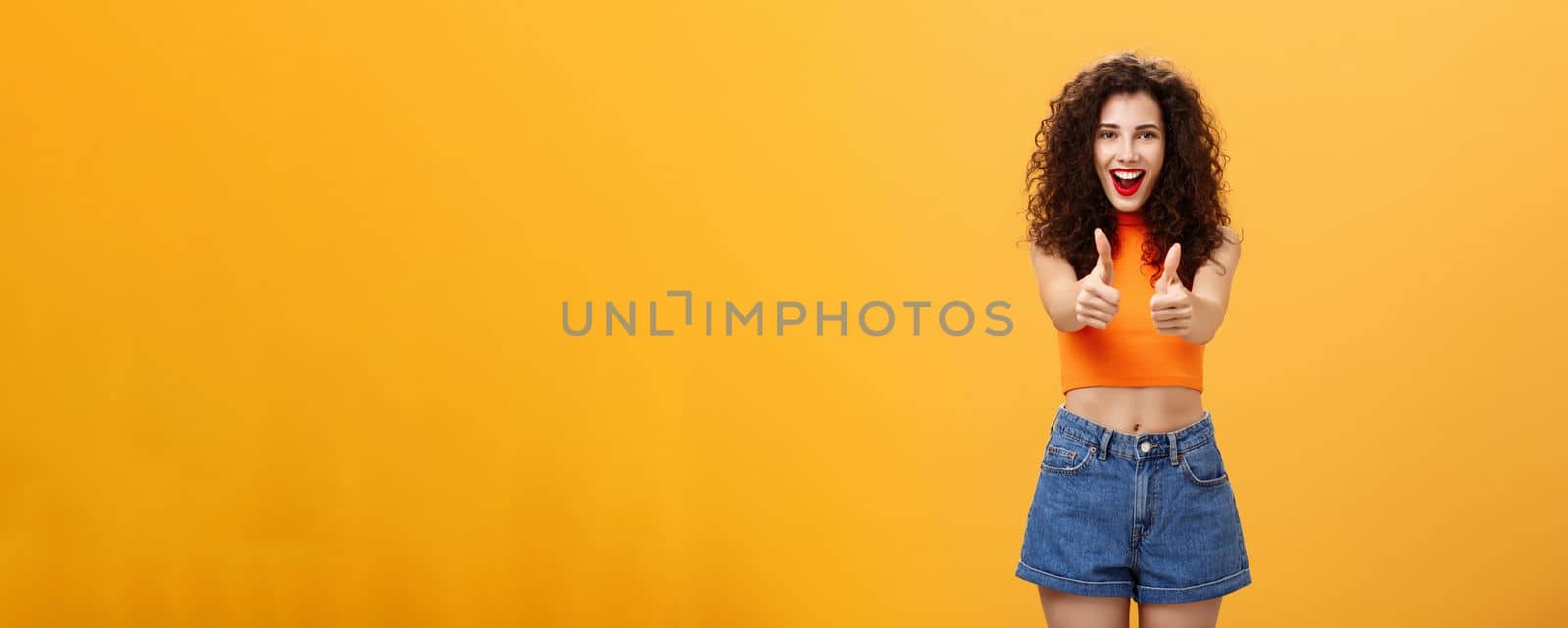 Charismatic ambitious and outgoing charming caucasian. woman with curly hairstyle and red lipstick showing thumbs up gesture in like or approval smiling joyfully being supportive over orange wall.