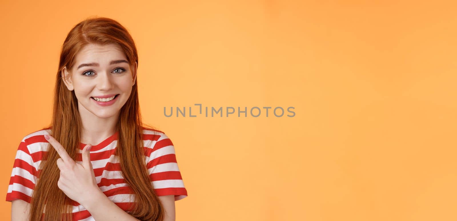 Close-up silly cute redhead female trying help showing way, smiling friendly politely introduce promo, pointing upper left corner smiling modest, stand orange background give advice.