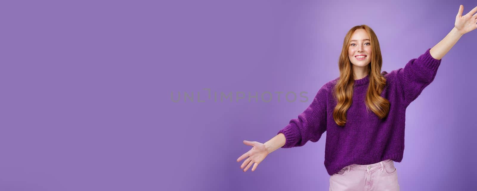 Girl stretched hands sideways to welcome and greet friend giving warm hug smiling broadly at camera standing joyful wanting cuddle over purple background, wearing violet sweater and pants. Copy space