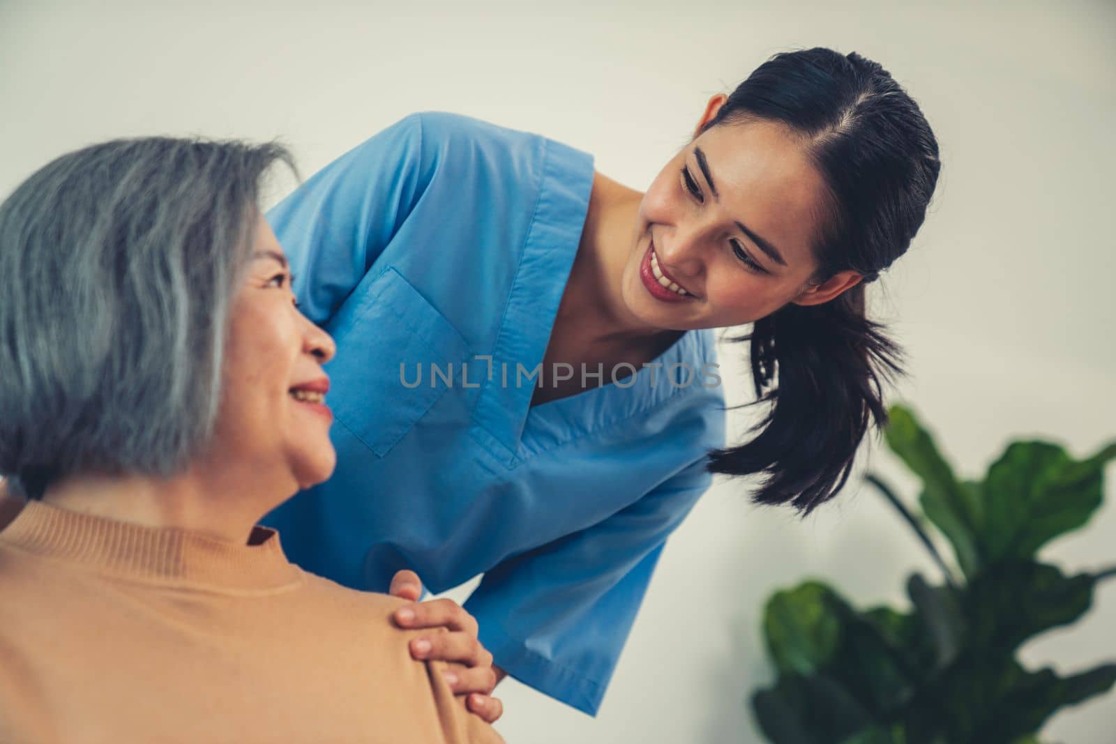 A caregiver rest her hands on the shoulders of a contented senior patient while she sitting on the sofa at home.