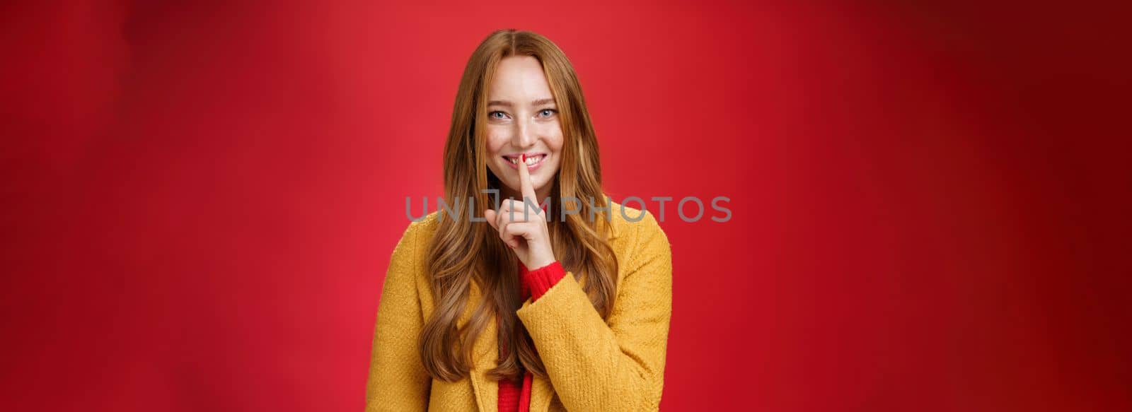 Shh keep it secret. Flirty and gentle attractive ginger girl with freckles teeling mysterious things showing shush gesture with index finger on mouth, smiling having surprise over red background.