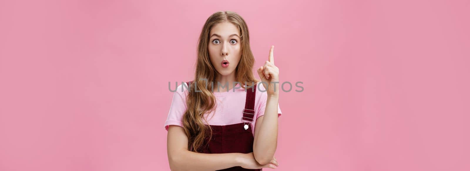 Young smart and creative female student sharing with team awesome idea coming in mind raising index finger in eureka gesture looking excited at camera folding lips saying suggestion over pink wall. Body language, body-positive and gestures concept