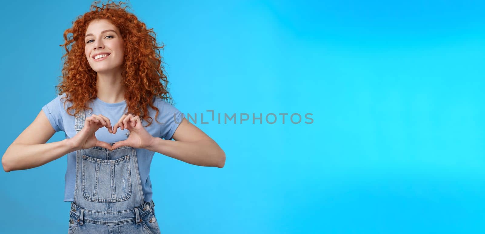 Sassy confident good-looking modern redhead curly woman raise head proud love tell girlfriend romantic heartwarming feelings present own heart smiling broadly standing blue background.