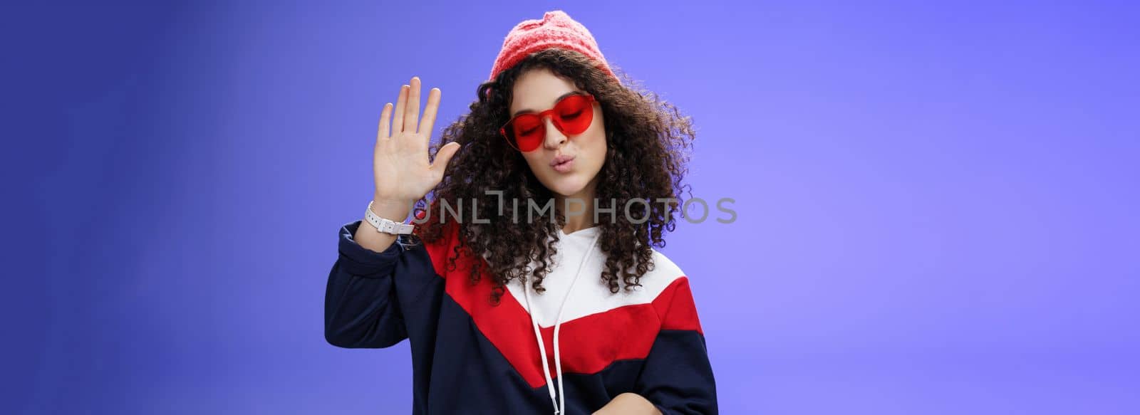Studio shot of cool and stylish dj girl in red beanie and sunglasses raising palm as enjoying cool music and dancing to rhythm folding lips having fun at awesome party posing chill over blue wall by Benzoix