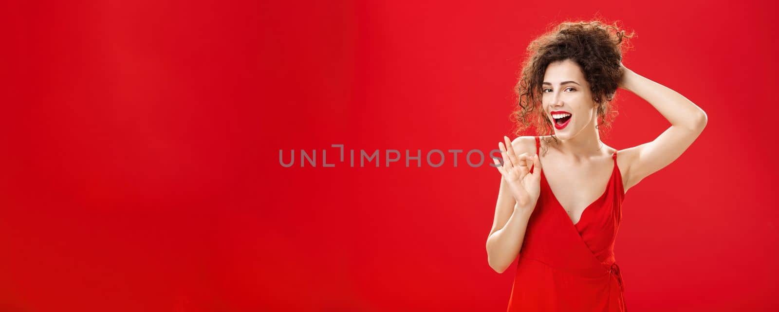 Portrait of sensual and sexy pleased carefree adult woman. gathering hair on back with hand smiling joyfully and confident while showing okay or approval gesture smiling assuring over red background.