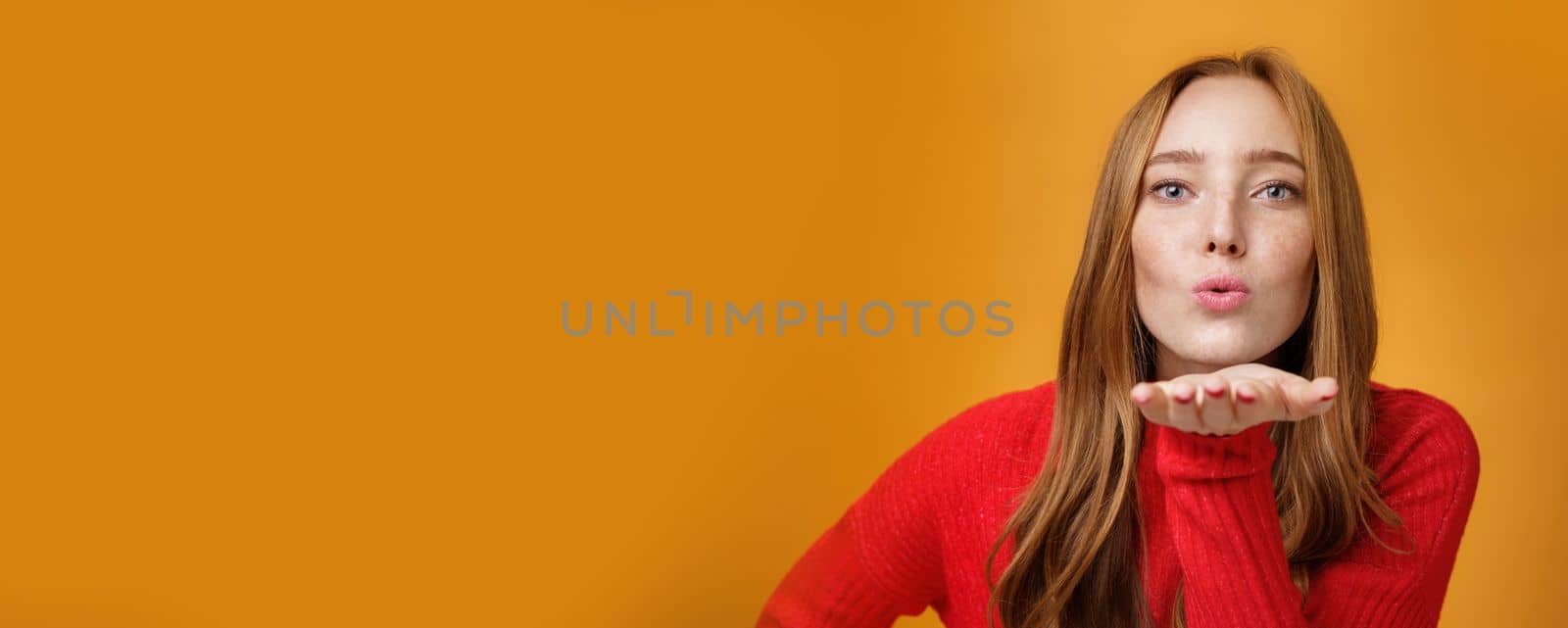 Lifestyle. Close-up shot of sensual and attractive redhead female in red knitted outfit folding lips tilting forward as sending air kiss at camera giving mwah holding palm near mouth over orange wall.