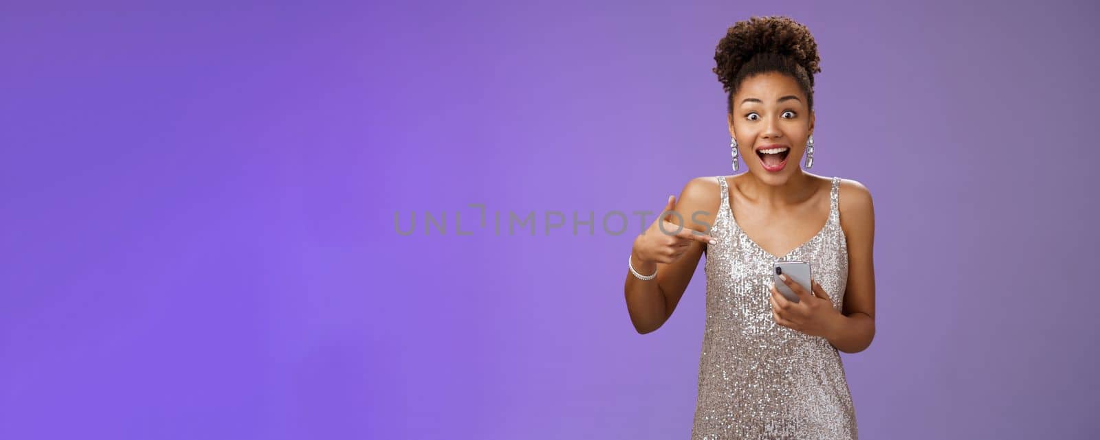 Amused charming glamour african-american woman in glittering silver dress astonished thrilled pointing smartphone receive incredible good perfect news telling you, standing blue background.