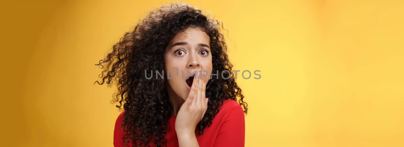 Close-up shot of concerned and shocked worried woman with curly hairstyle open mouth wide and covering it with palm popping eyes at camera impressed and upset over yellow background. Emotions and faciale xpressions concept