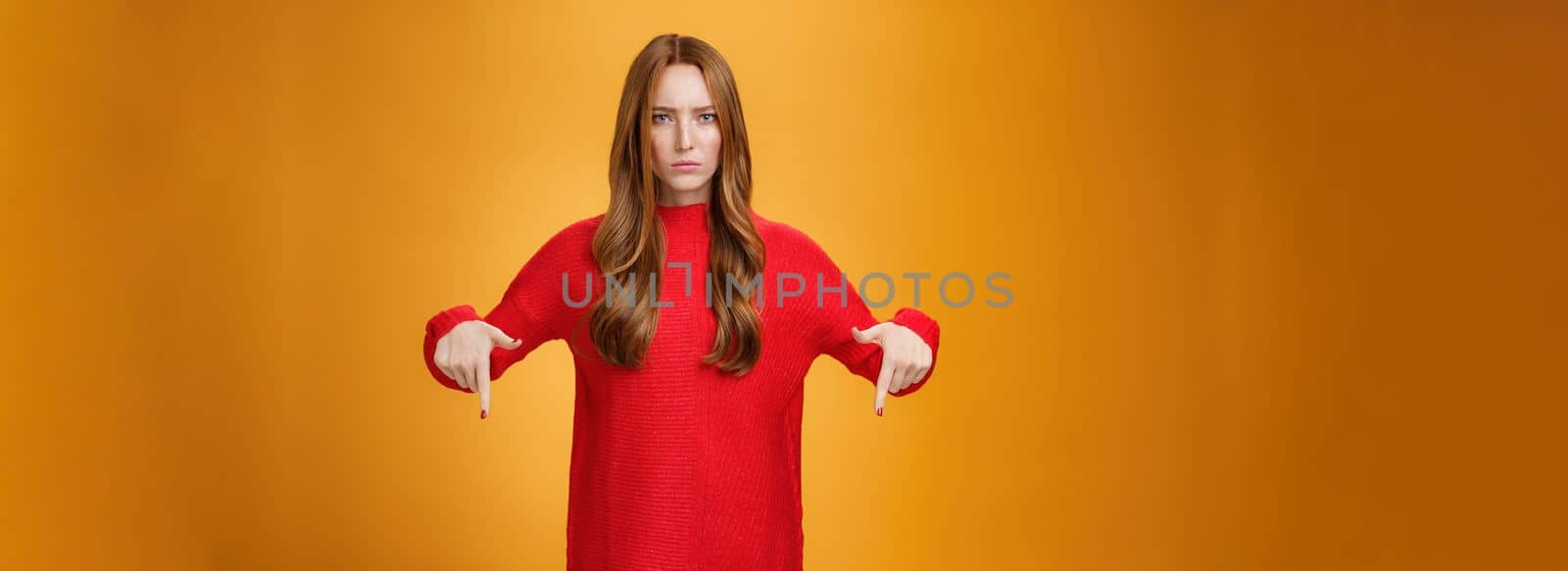 Displeased serious-looking female in knitted red dress frowning looking concerned as pointing down at displeasing unappropriate thing standing disappointed and unhappy over orange background.