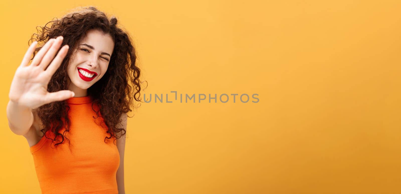 Give me five mate. Stylish friendly and joyful attractive woman with curly hairstyle winking and smiling happily pulling hand towards camera to greet or congratulate friend over orange background. Copy space