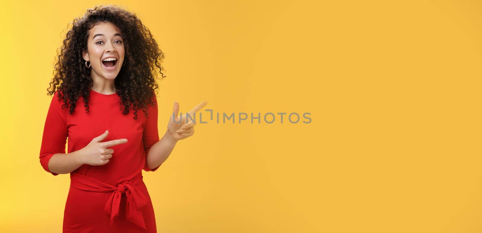 Thrilled and happy joyful young charismatic woman with curly hair in red dress laughing out loud and smiling amazed, delighted pointing at upper right corner impressed over yellow background.