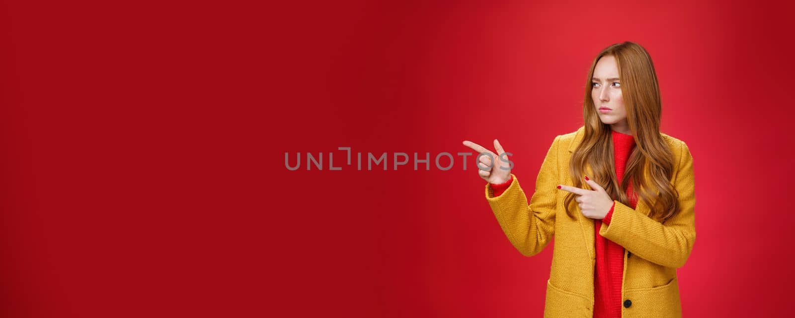 Studio shot of intense suspicious and displeased redhead female in yellow coat pointing and looking serious left squnting and frowning making focused look at something disturbing and doubtful. Emotions and facial expressions concept