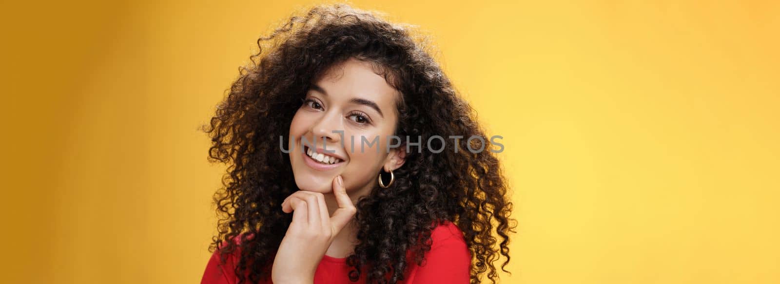 Lifestyle. Headshot of cute silly and tender feminine romantic woman with curly hairstyle touching lip with index finger making eyes at camera and smiling as using seduction skills over yellow background.