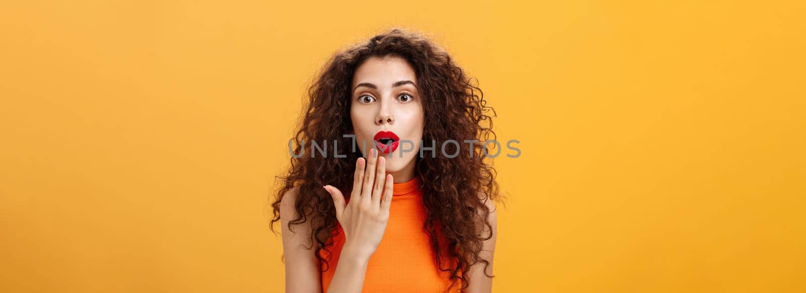 Waist-up shot of amazed and excited caucasian woman. with curly hair in red lipstic opening mouth from surprise covering it with palm listening shocking rumors reacting astonished to amusing news.