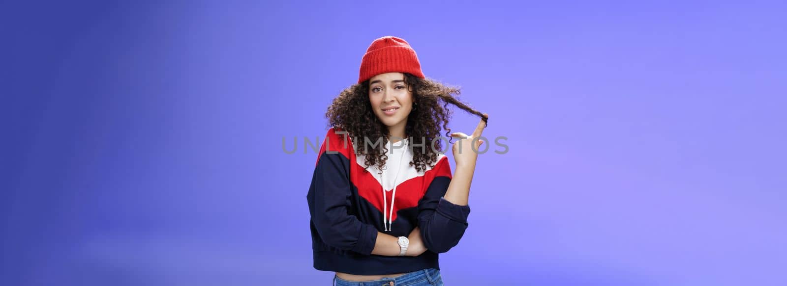 Stylish and snobbish arrogant curly-haired woman in warm beanie rolling curl on finger smirking and looking with contempt at camera, scorning person as being too cool over blue background.