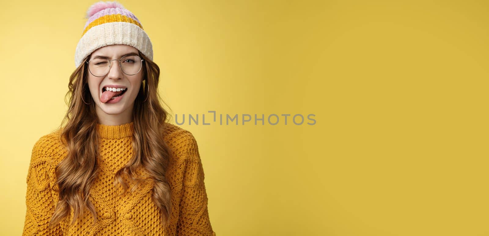 Carefree attractive funny happy caucasian girl 20s curly-haired wear glasses hat sweater having fun playfully show tongue, winking flirty expressing positive optimistic mood, yellow background.