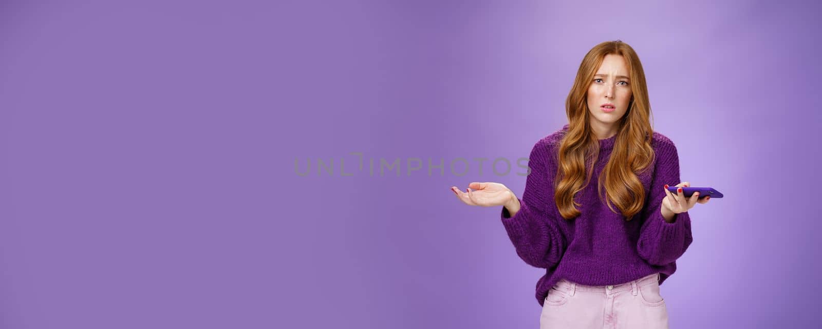 Clueless and sad cute redhead woman spread hands sideways frowning disturbed and upset holding smartphone being questioned and clueless why mobile phone not working, posing over purple wall.