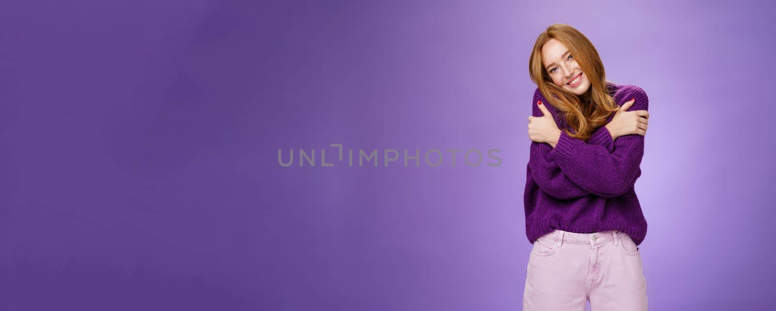 Lifestyle. Silly and cute carefree redhead woman in 20s leaning on shoulder as hugging herself feeling warmth wearing purple sweater smiling broadly in cozy and relaxing atmosphere over violet wall.