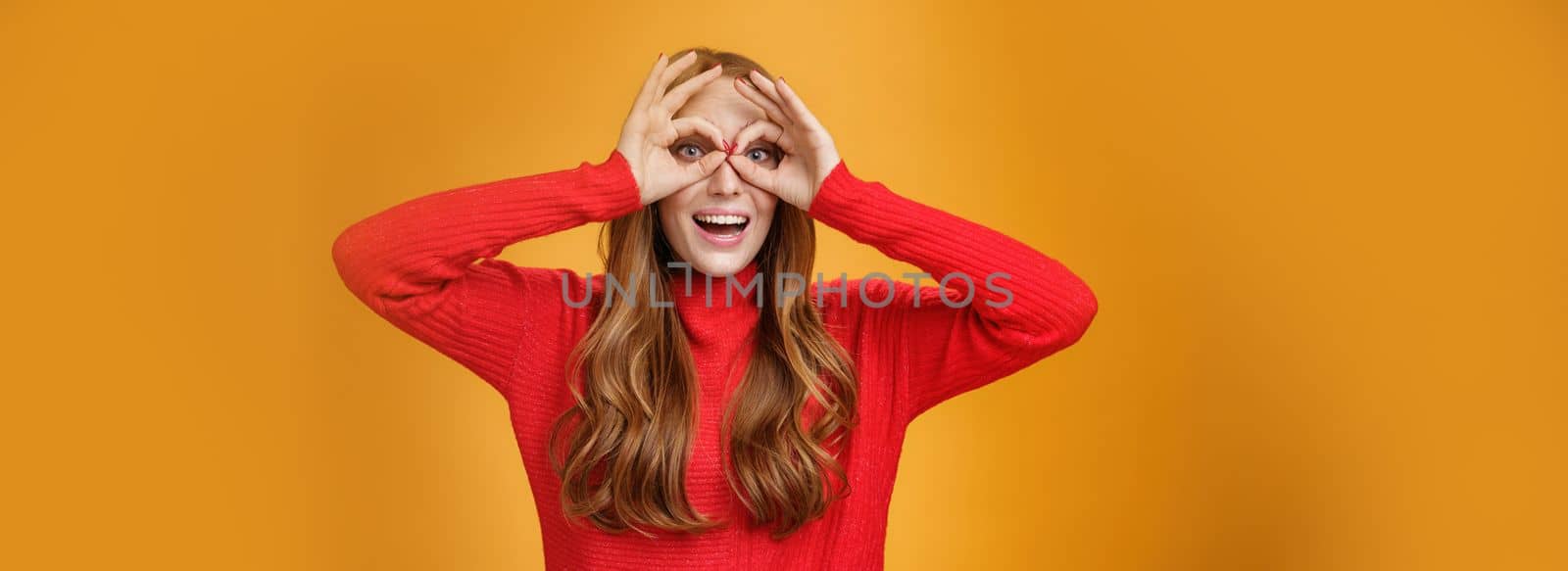 Waist-up shot of childish and playful funny ginger girl in red knitted dress making faces showing circles over eyes like goggles and smiling broadly having fun against orange backgound like child. Copy space