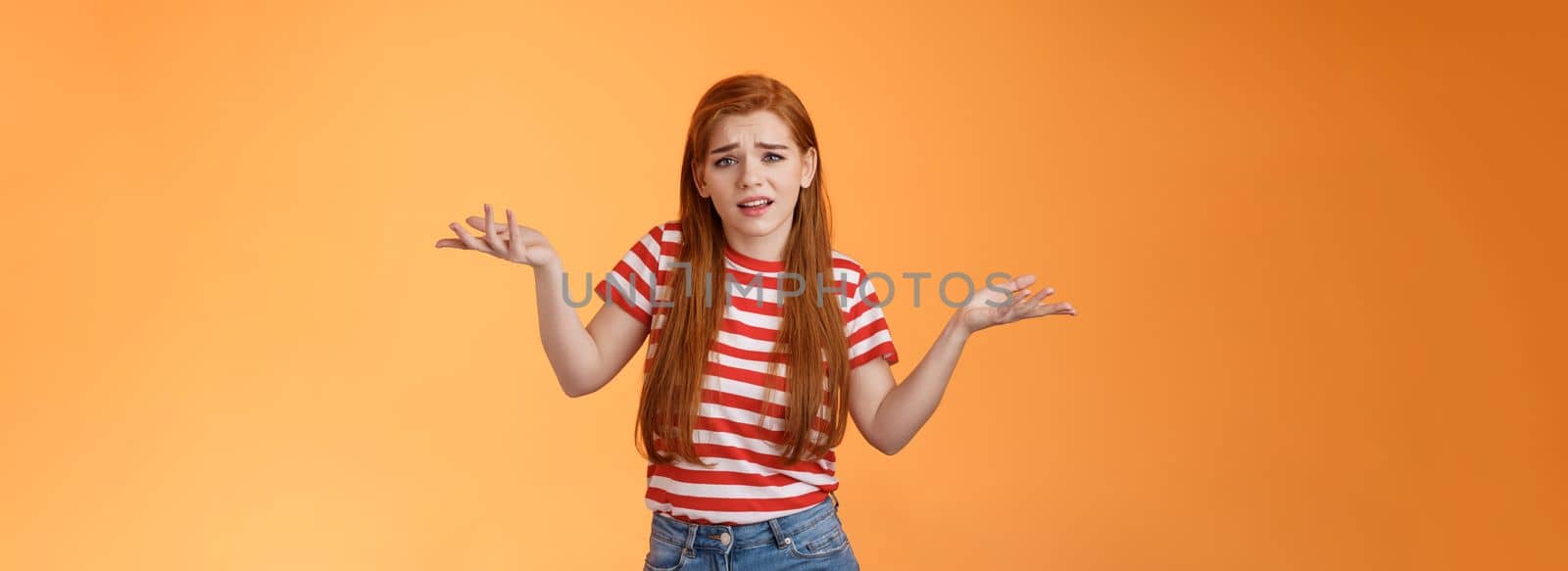 Careless arrogant redhead rich girl say so what, not giving care what people thing, look dismay ignorant shrugging hands spread sideways, uninterested, give no concern, stand orange background.