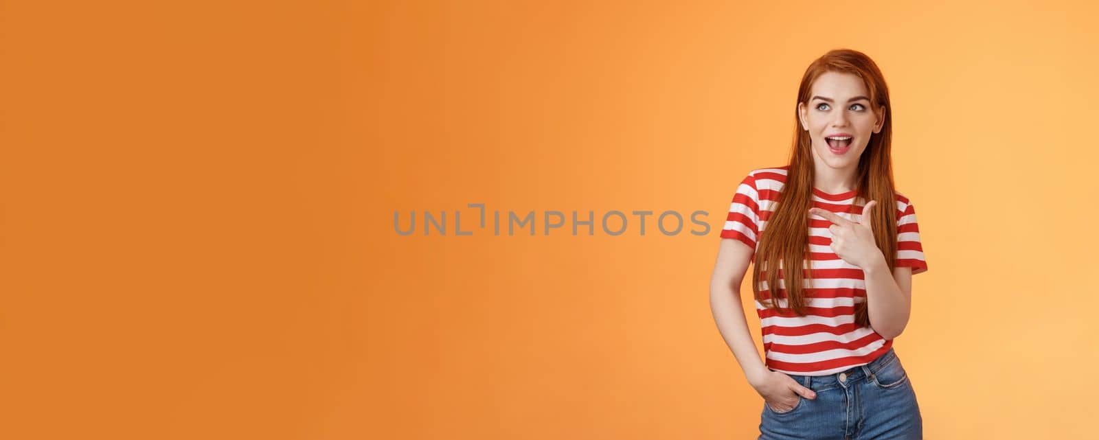 Enthusiastic amazed sassy modern redhead female attend impressive bad awesome design, look amused, observe cool place, pointing left excited, open mouth fascinated intrigued, stand orange background.