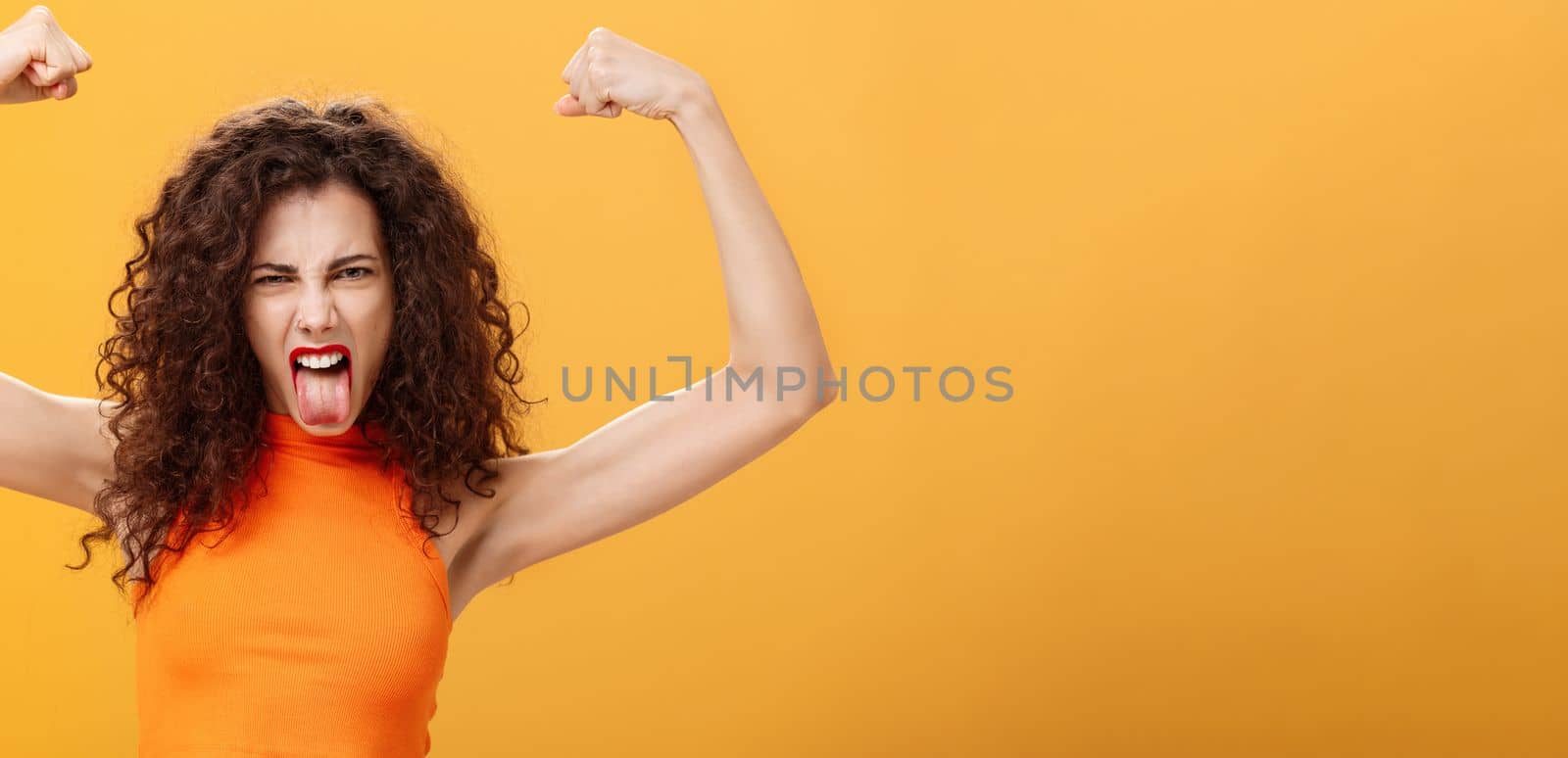 Waist-up shot of cool and daring caucasian female in orange top with tattoo on arm frowning making funny face sticking out tongue raising hands showing muscles feeling power and strengths. Copy space