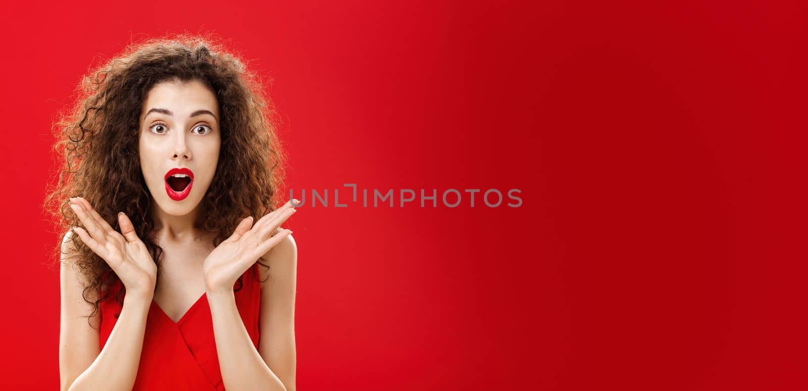 Waist-up shot of amused and astonished good-looking young elegant female. with curly hairstyle in red lipstick raising palms in surprise opening mouth and looking at camera intrigued and shocked.