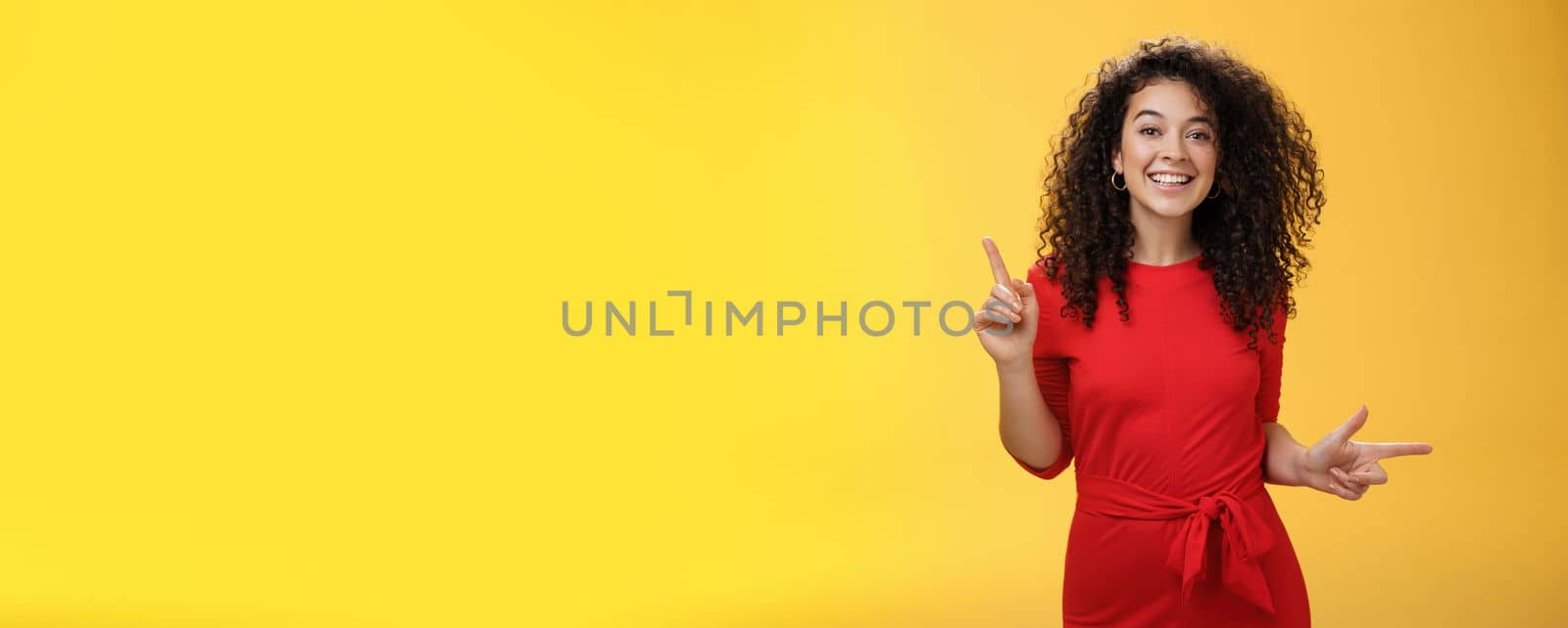 Charismatic cute and happy young curly-haired european female in red dress dancing and pointing up and right, showing sideways as presenting choices picking, making decision and smiling at camera.