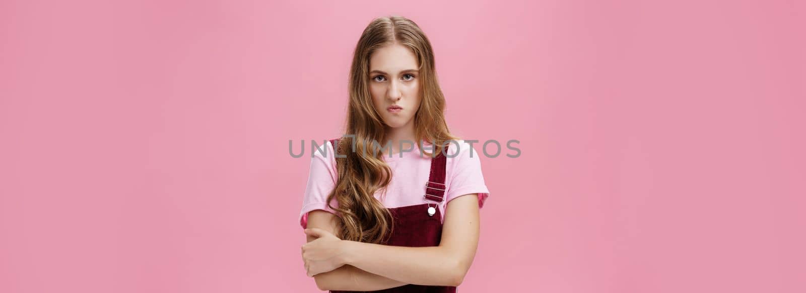 I mad on world. Unhappy hateful young arrogant girl in overalls with wavy natural hairstyle crossing arms against chest in defensive pose lifting upper lip in scorn and disdain, looking with anger. Emotions concept