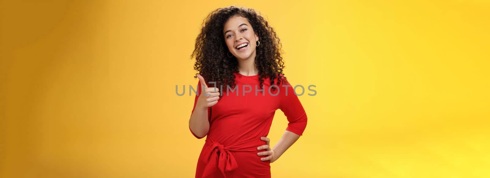 Portrait of happy optimistic young curly-haired woman in red dress laughing joyfully, showing thumbs up in approval and like gesture, delighted with awesome idea, accepting plan over yellow wall by Benzoix