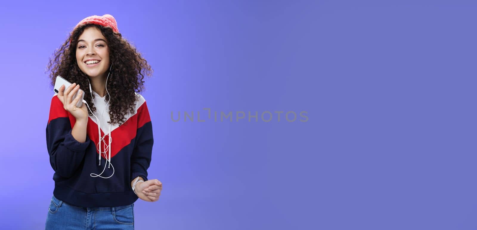 Waist-up shot of charming happy tender curly-haired female in beanie and sweatshirt listening music as dancing carefree, playing karaoke app or enjoying cool song in earphones over blue background.
