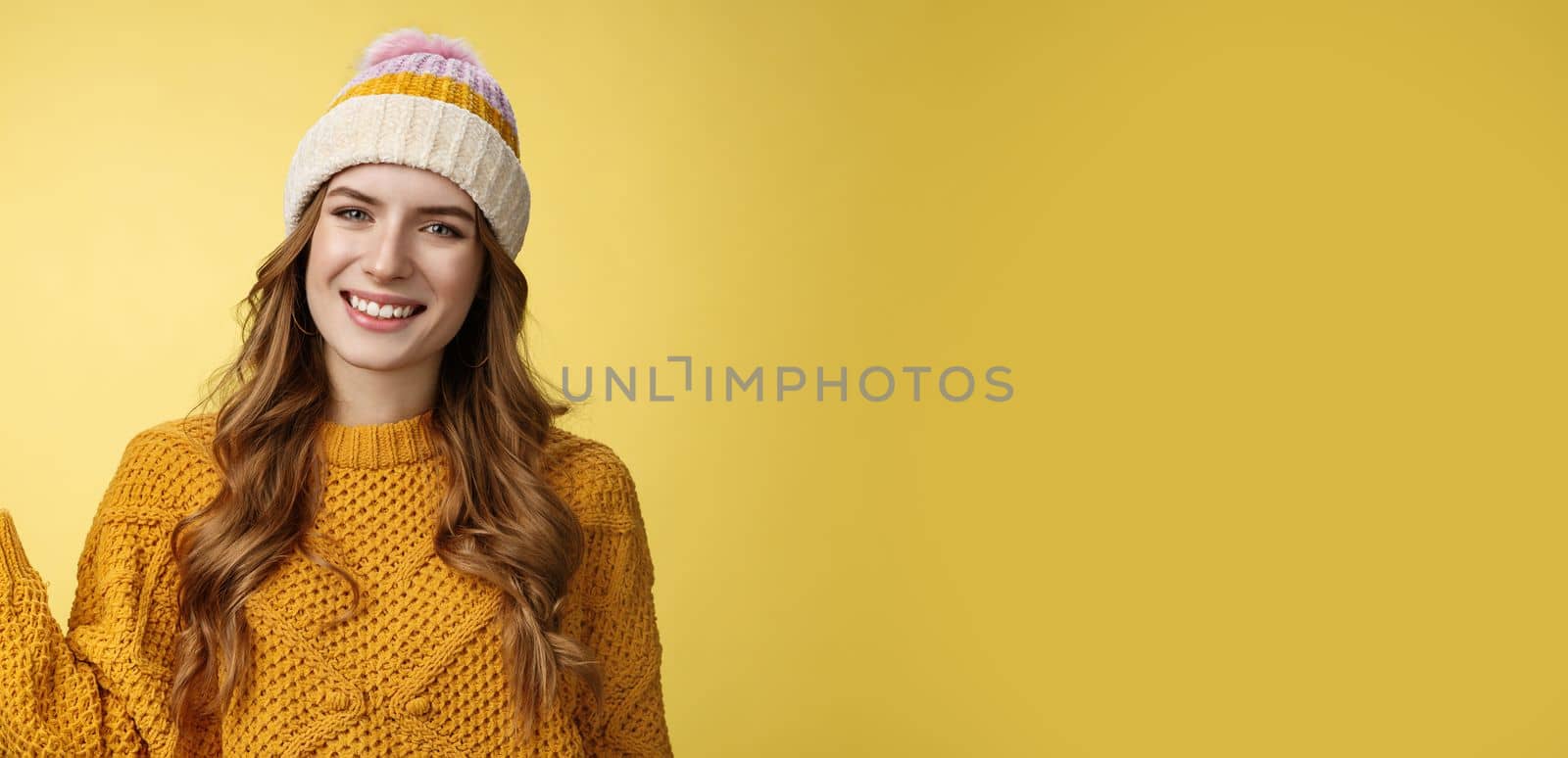 Friendly-looking confident pleasant attactive young european woman wearing winter hat, sweater extend arm show product welcoming inviting guests come in smiling happily, promoting copy space.