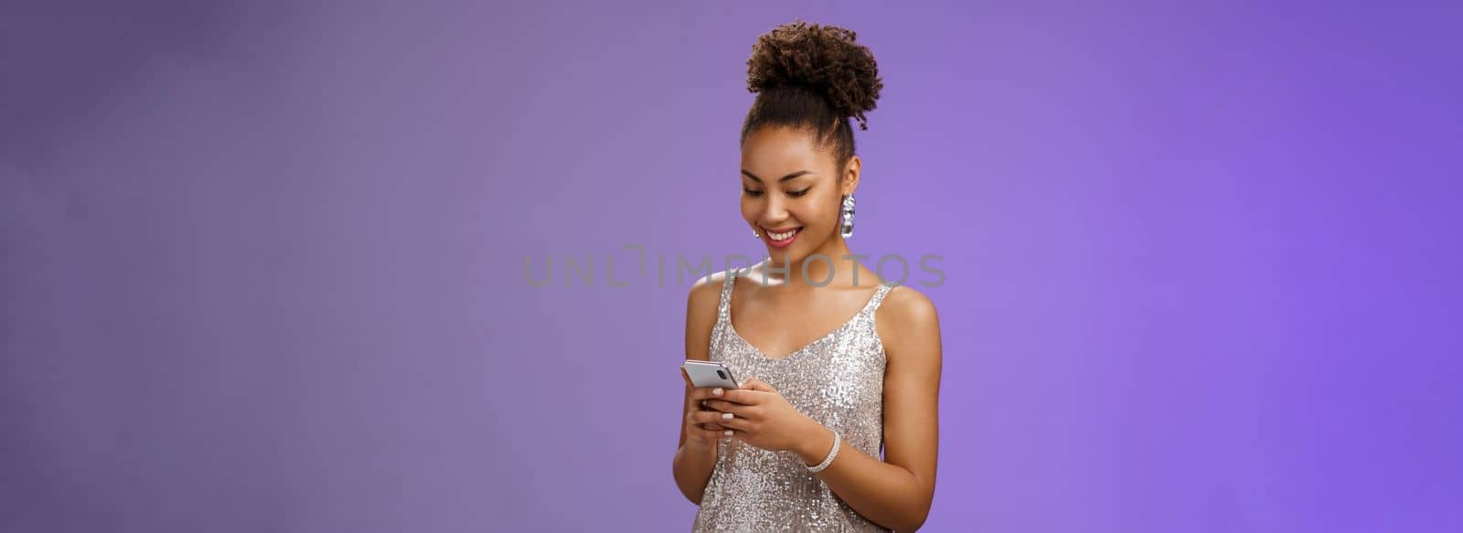 Charming elegant african-american woman in stylish glittering prom dress writing message calling friend hold smartphone look amused happy smile phone display using gadget app scroll party photos by Benzoix