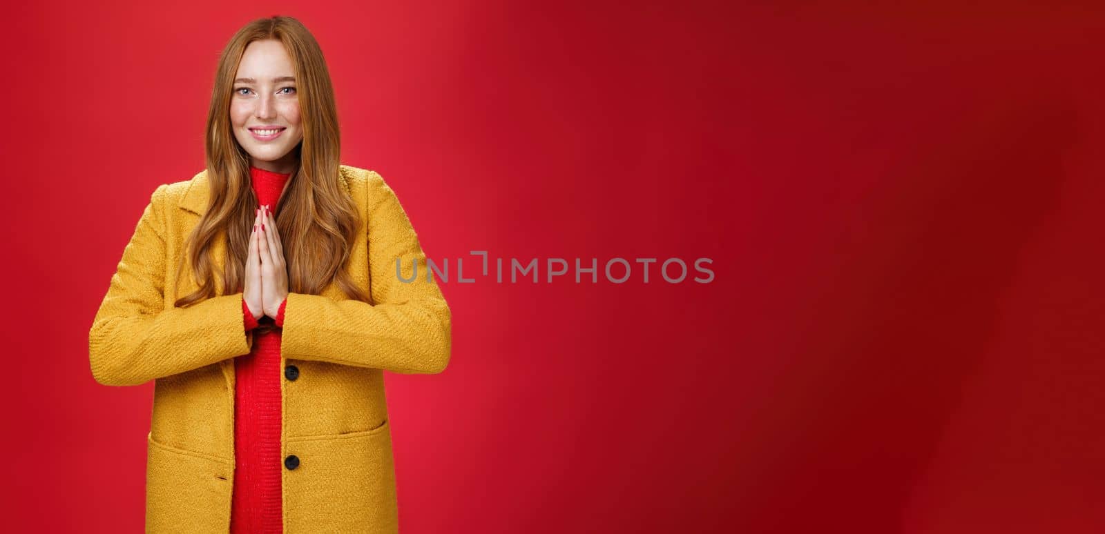 Girl welcomes us with buddhist namaste gesture pressing palms together over chest smiling friendly and delighted with carefree emotions holding hands in pray, posing happily over red background.
