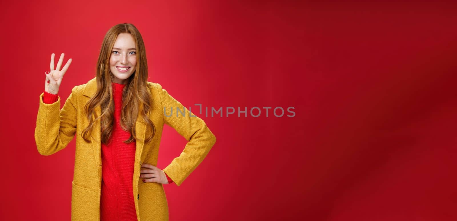 Cute friendly-looking ginger girl showing three reasons buy product smiling broadly making third reservation, looking delighted and confident in her choice posing against red background in yellow coat.