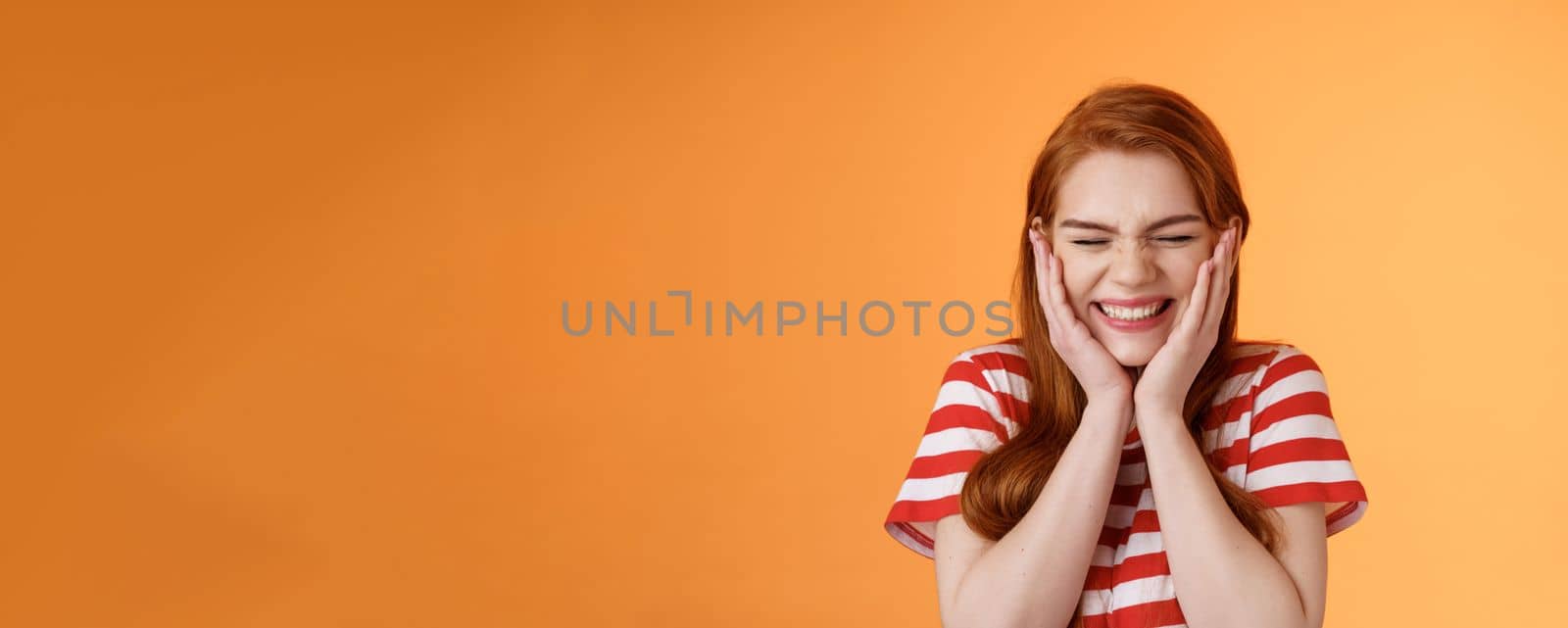 Close-up cheerful lucky excited smiling girl, cannot believe cheering hear excellent news, close eyes celebrating, touch cheeks blushing joy delight, rejoicing awesome situation, orange background.