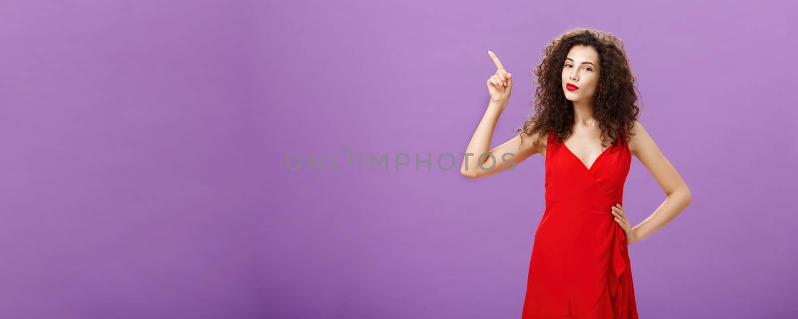 Woman ready to shine on dance floor in elegant red dress with sensual makeup and curly hairstyle pointing at upper left corner smirking with delight and self-assured expression over purple wall. Advertisement concept