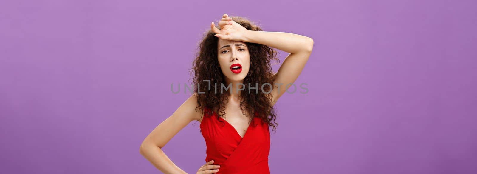 Woman fainting feeling bad whiping sweat of forehead standing drained and exhausted over purple background in red stylish dress expressing gloomy and unhappy feelings wanting some help. Copy space