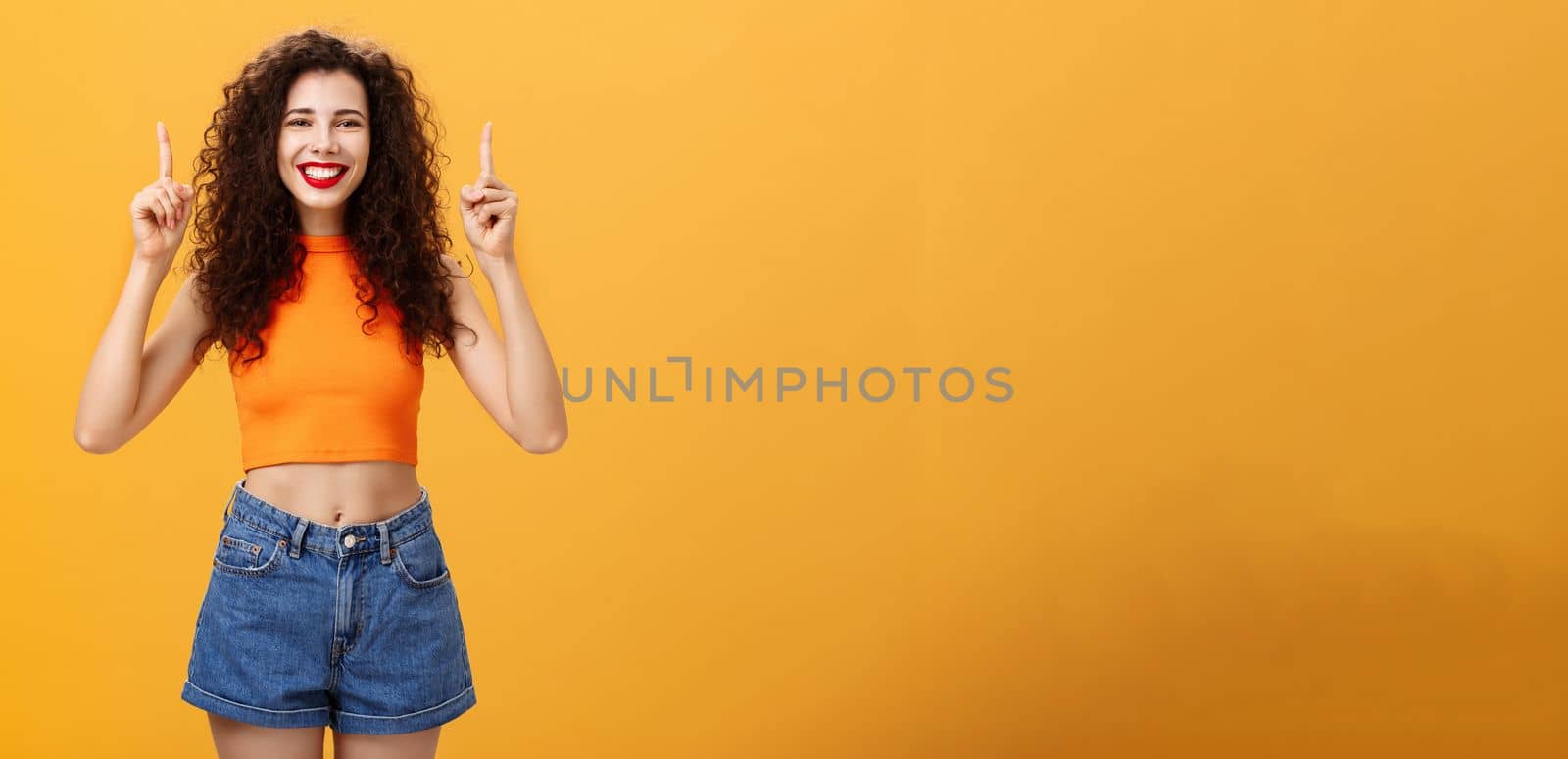Indoor shot of friendly and cute happy relaxed female with red lipstick and curly hairstyle wearing cool cropped top and denim shorts smiling broadly pointing up posing over orange background. Copy space