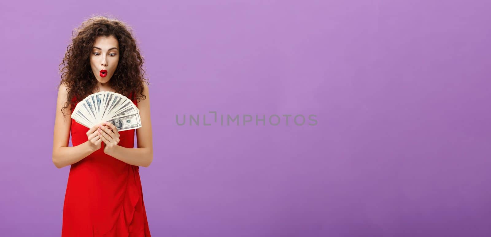 Portrait of surprised and amazed speechless woman folding lips making wow sound while holding and looking at lots of money being rich and successful receiving lots of cash over purple background. Finance concept