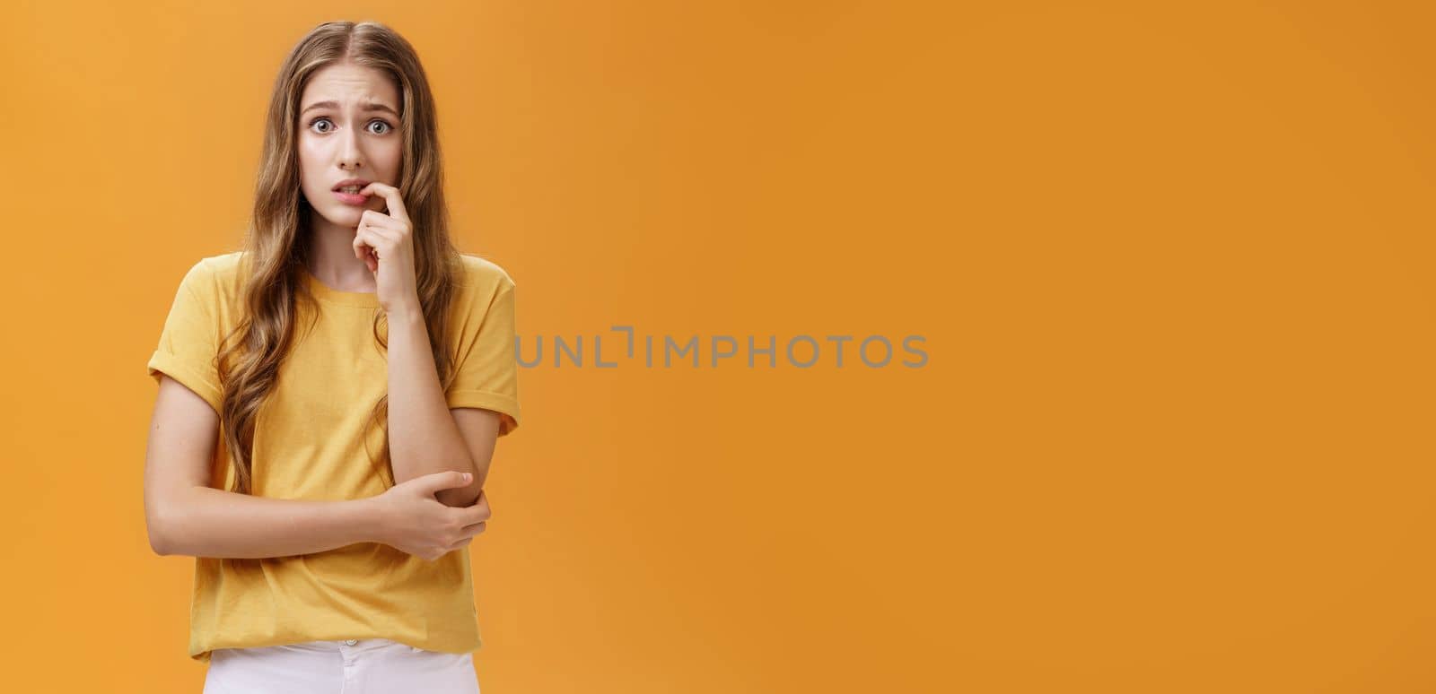 Girl worried mom react on new tattoo. Portrait of anxious and nervous silly insecure young female with wavy natural hairstyle biting finger panicking looking concerned and troubled over orange wall. Emotions concept