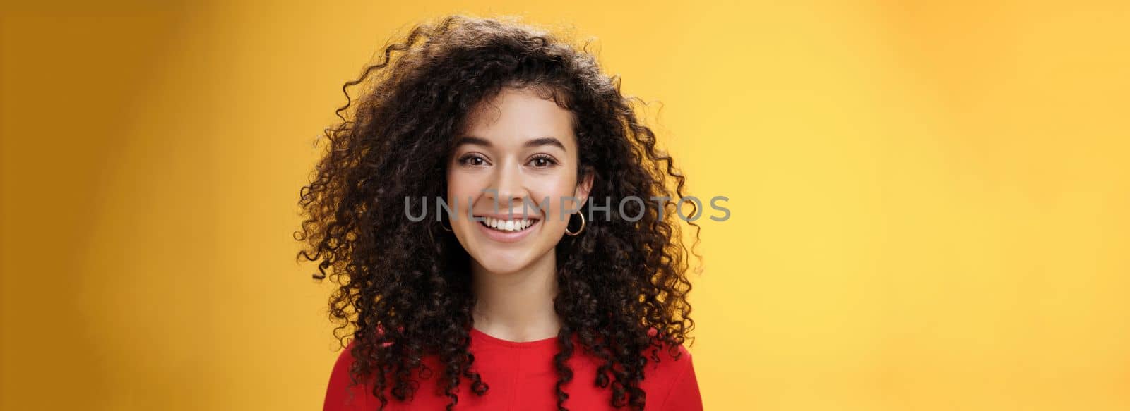 Close-up shot of pretty caucasian girl with curly hair in red dress and earrings smiling joyfully with pleased hopeful expression gazing at camera carefree, posing over yellow background.