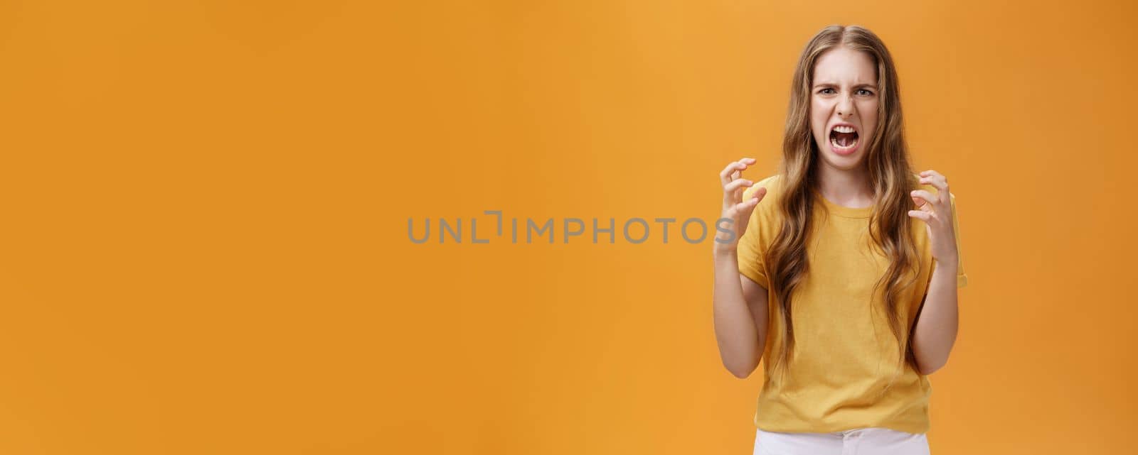 Portrait of irritated cute european female squeezing hands in fists with anger frowning and making hateful face standing annoyed and pissed over orange background with furious look. Emotions concept